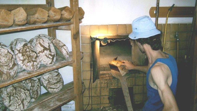 A baker in a blue tank top and hat takes bread out of an oven. Shelves with loaves of bread can be seen in the background.