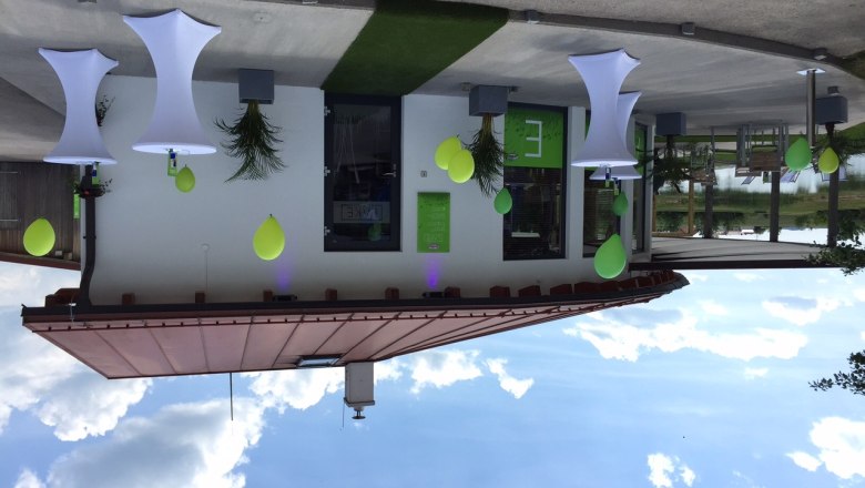 Eine moderne Strandbar mit weißen Stehtischen und grünen Dekorationen vor einem Gebäude mit Flachdach.