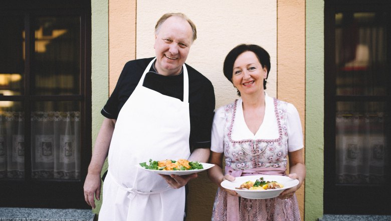 A man in an apron and a woman in traditional dress hold plates of food in front of a building.