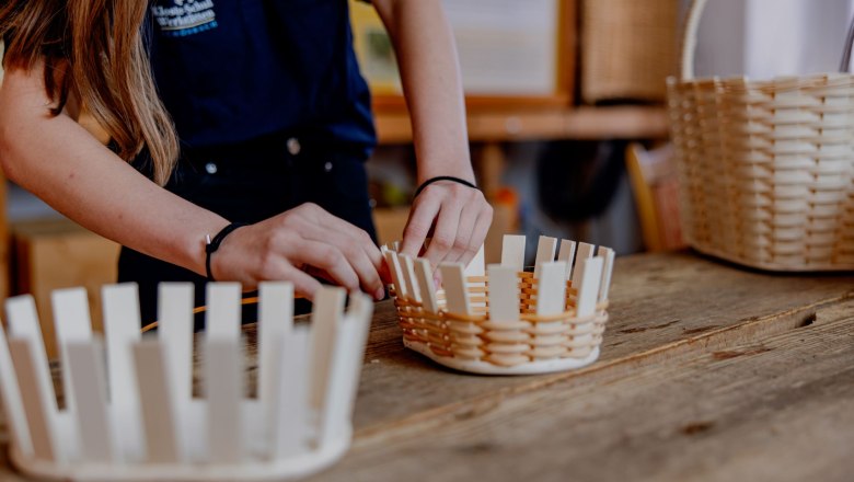 Person flechtet einen Korb aus Holzstreifen auf einem Holztisch.