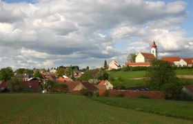 Landschaft mit Dorf und Kirche unter bewölktem Himmel.
