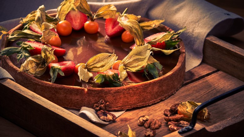 A cake with chocolate icing, decorated with strawberries and physalis, on a wooden tray.
