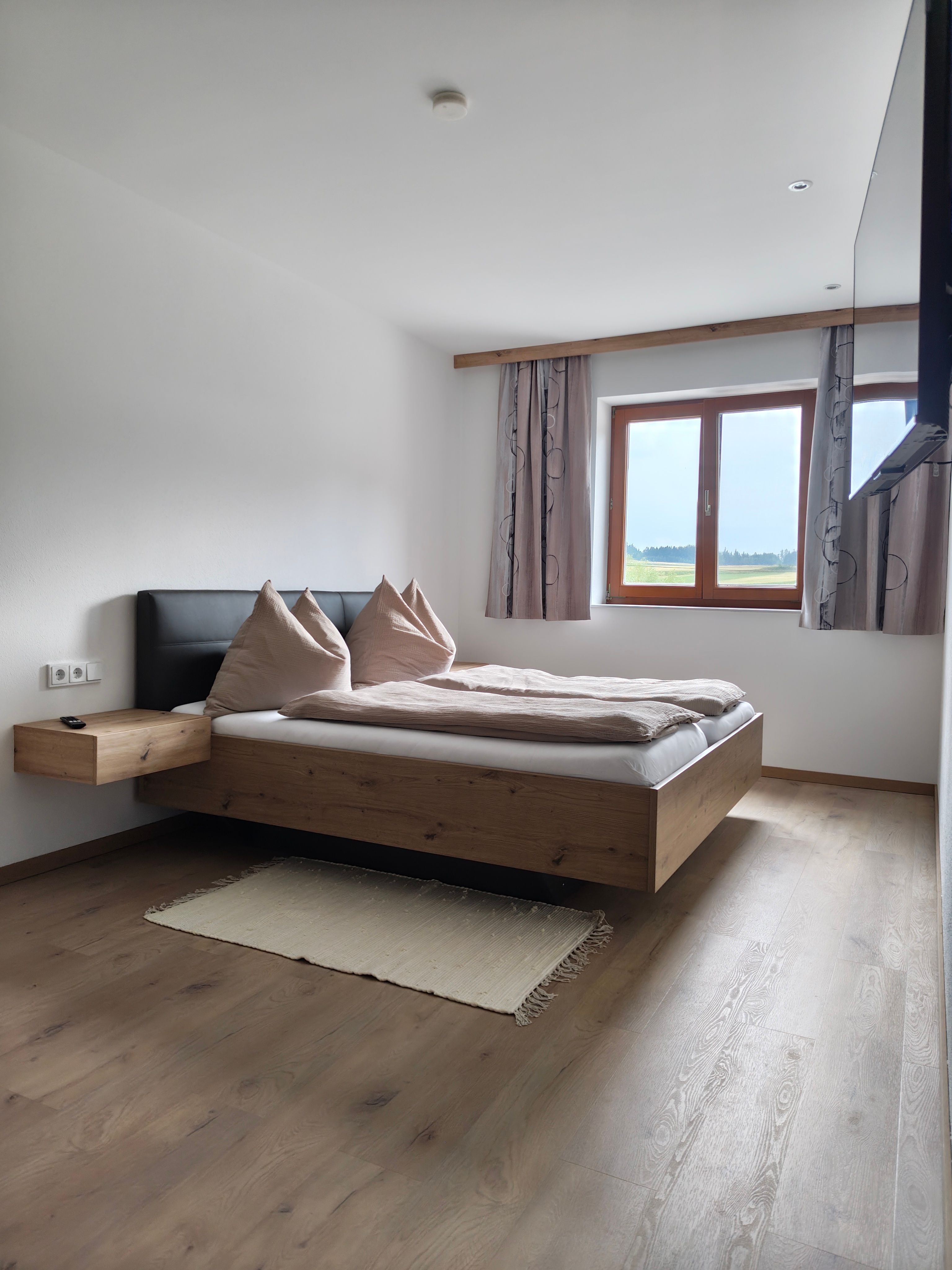 Modernes Schlafzimmer mit Holzbett, Kissen, Fenster mit Vorhängen und Blick ins Grüne.