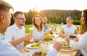 Menschen stoßen bei einem Essen im Freien an, umgeben von Natur.