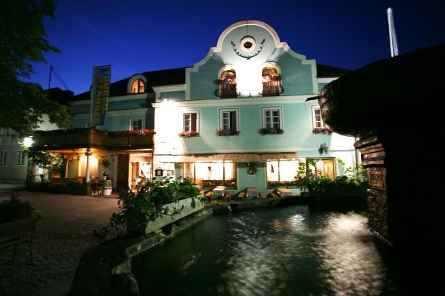 Ein traditionelles Gasthaus mit blauer Fassade und beleuchteten Fenstern bei Nacht, im Vordergrund ein Brunnen.