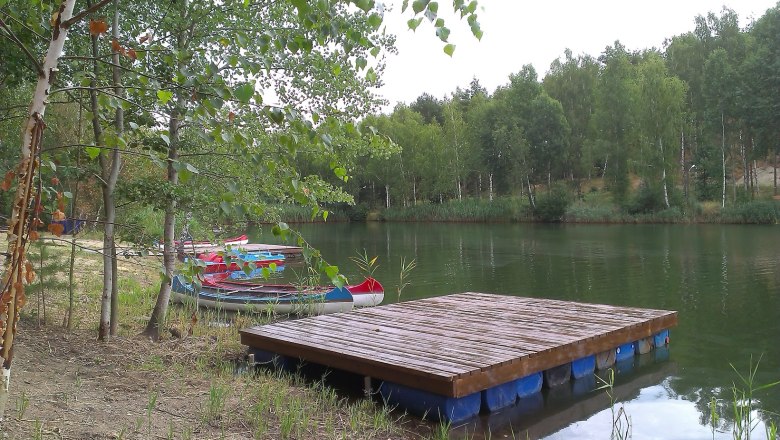 Ein ruhiger See mit einem Holzsteg und mehreren Kanus am Ufer, umgeben von Bäumen.