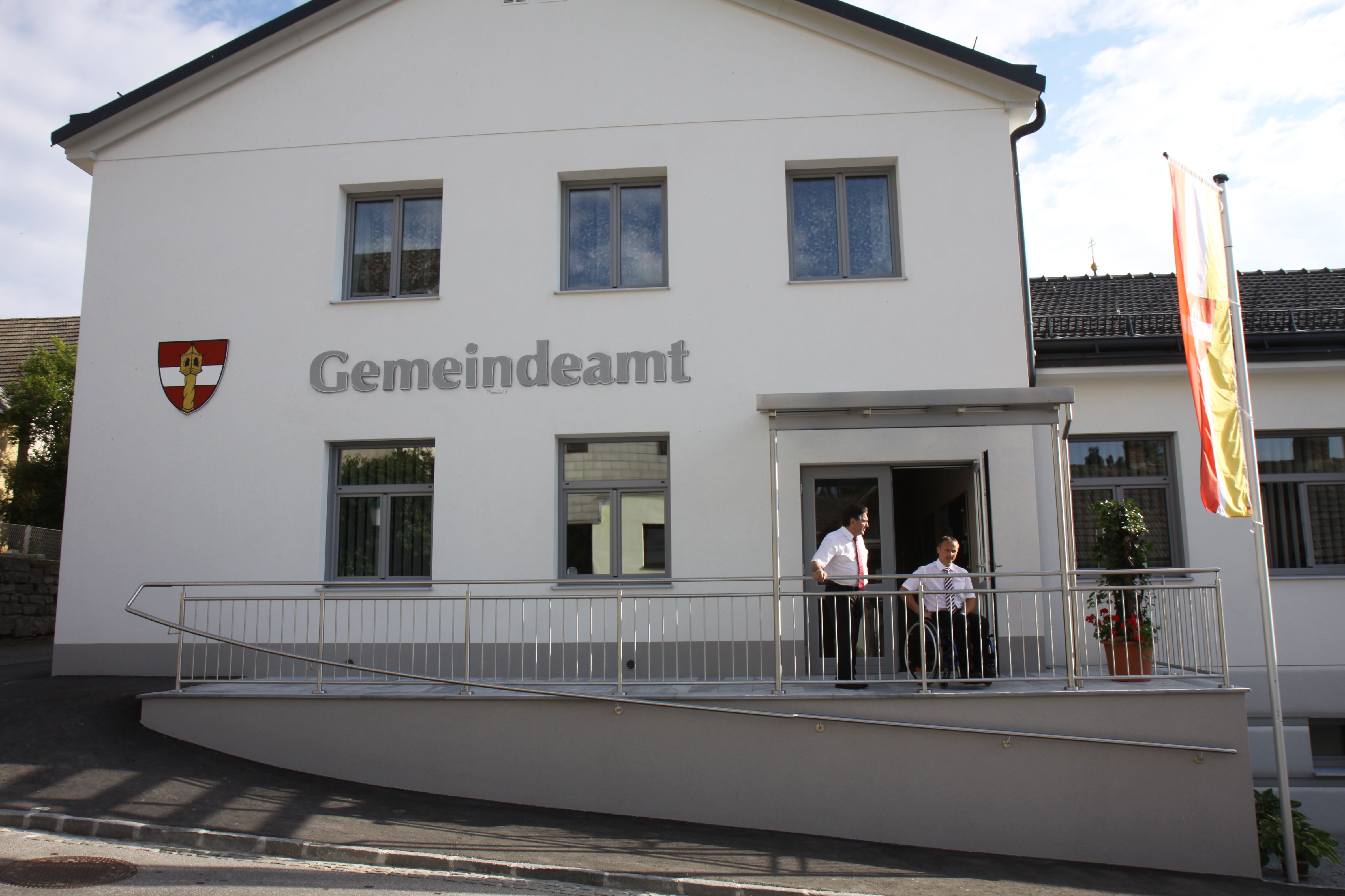 Ein modernes Gemeindeamt mit weißer Fassade, zwei Personen stehen auf einer Rampe, daneben weht eine Flagge.