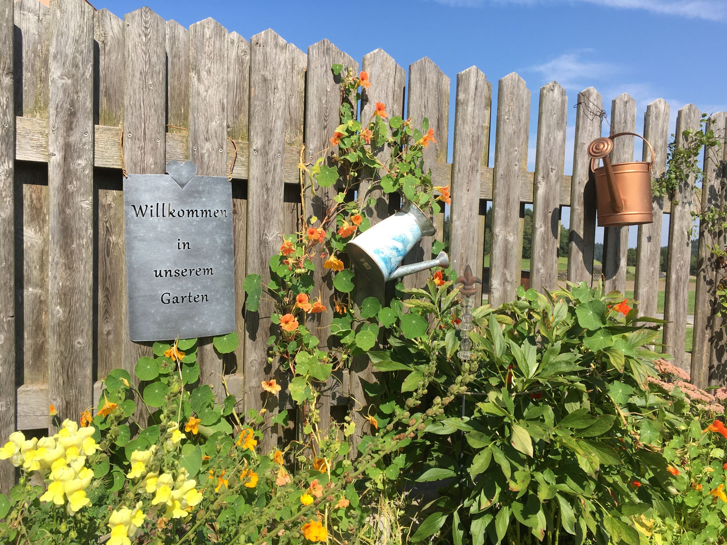 Ein Holzzaun mit einem Schild 'Willkommen in unserem Garten', umgeben von Blumen und Gießkannen.