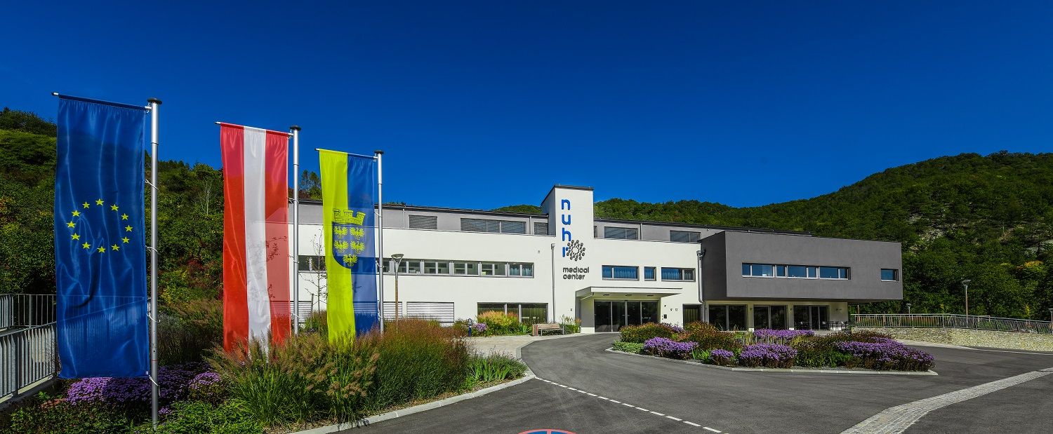 Modernes Gebäude mit drei Flaggen davor, umgeben von grüner Landschaft.