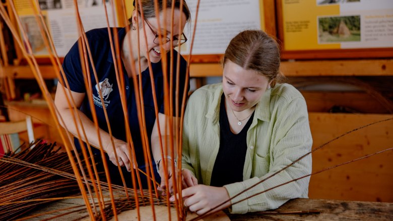 Zwei Frauen flechten gemeinsam Weidenruten an einem Tisch.