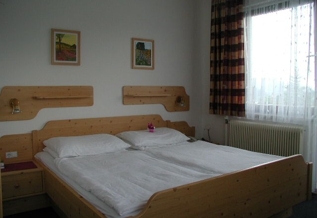 Ein einfach eingerichtetes Hotelzimmer mit einem Doppelbett aus Holz, zwei Bildern an der Wand und einem Fenster mit Vorhängen.