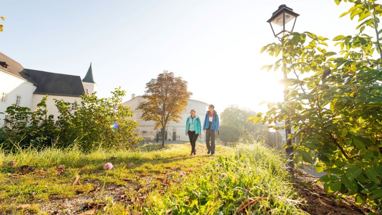 Zwei Personen spazieren auf einem sonnigen Weg neben einem Gebäude mit Turm.