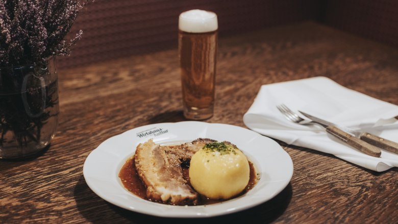 Ein Teller mit Schweinsbraten und Waldviertler Knödel, daneben ein Glas Bier auf einem Holztisch.