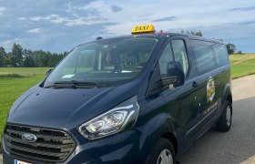 Ein blaues Ford-Taxi steht auf einer Landstraße vor einer grünen Wiese und einem bewölkten Himmel.