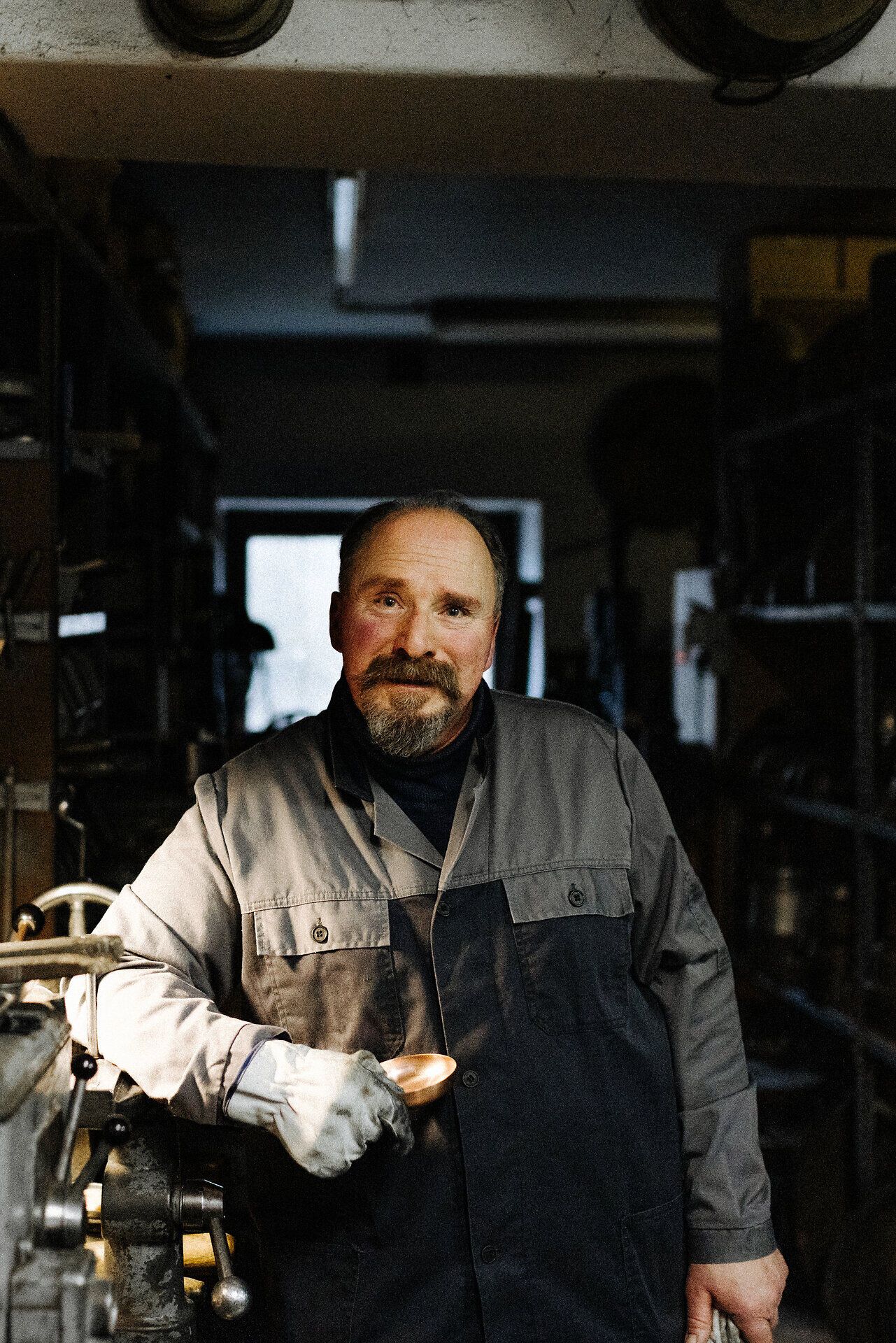 In einer warmen, einladenden Werkstatt strahlt der Handwerker Stolz aus, während er eine glänzende Kaffeekanne in der Hand hält. Umgeben von Werkzeugen und Metallformen, vermittelt die Szene die Leidenschaft und das handwerkliche Können, das in jedes Stück einfließt. Hier wird Tradition lebendig und die Kunst des Metallverarbeitens zelebriert.