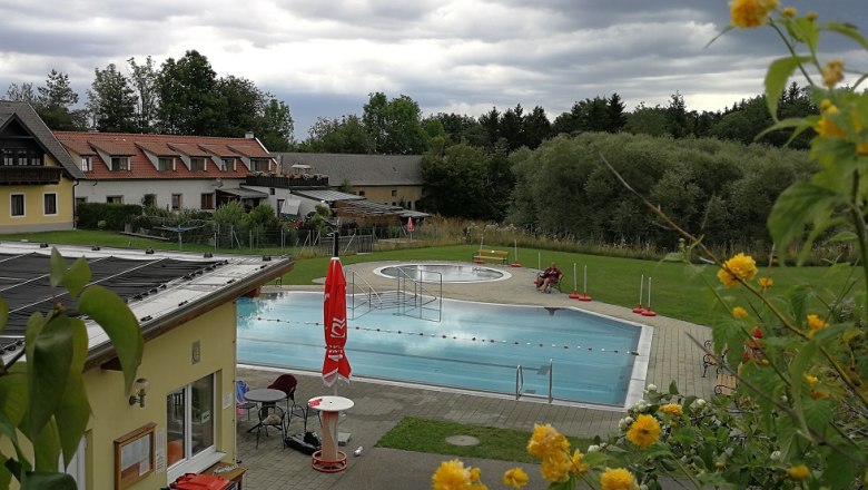 Ein Freibad mit Schwimmbecken, umgeben von Gebäuden und Bäumen, unter bewölktem Himmel.