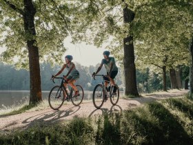 Zwei Personen, die man von vorne sieht, umgeben von Wald und Wasser, die mit ihren R&auml;dern fahren