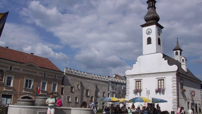 Ein Stadtplatz mit einem weißen Gebäude mit Turm und Uhr, Menschen sitzen unter Sonnenschirmen, ein Brunnen im Vordergrund.