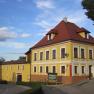 Yellow two-storey building with red roof and white window frames, next to it a smaller building. A church spire in the background.