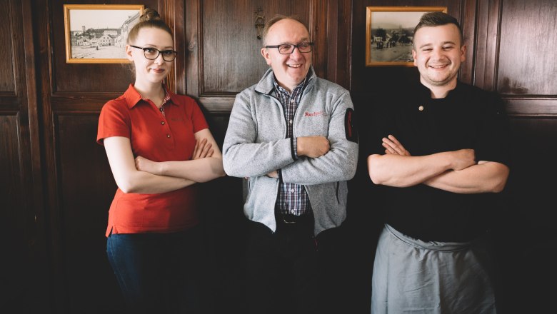 Three people stand smiling with their arms folded in front of a wooden wall.