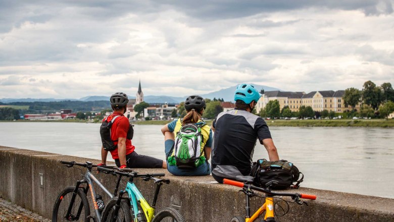 Drei Radfahrer sitzen auf einer Mauer am Flussufer und blicken auf eine Stadt mit Kirche und Gebäuden im Hintergrund.
