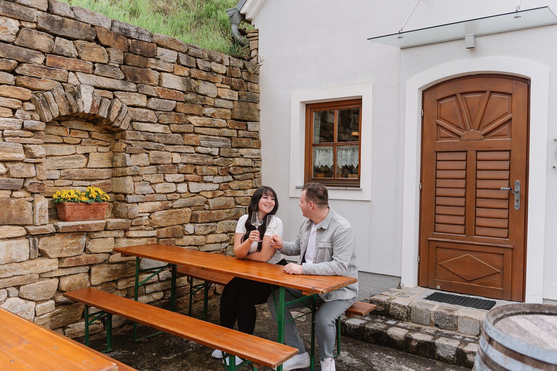 Inmitten der malerischen Weinlandschaft genießen zwei Menschen einen entspannten Moment an einem rustikalen Holztisch. Die sanften Hügel, bedeckt mit üppigen Weinreben, schaffen eine einladende Atmosphäre, die zum Verweilen einlädt. Hier, wo die Natur und die Kultur des Weins harmonisch verschmelzen, wird der Frühling zu einem unvergesslichen Erlebnis.