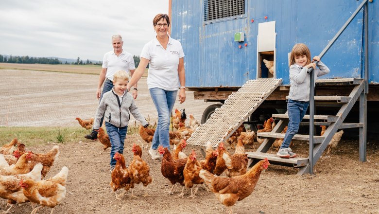 Familie mit Hühnern vor einem blauen Hühnerstall auf einem Feld.