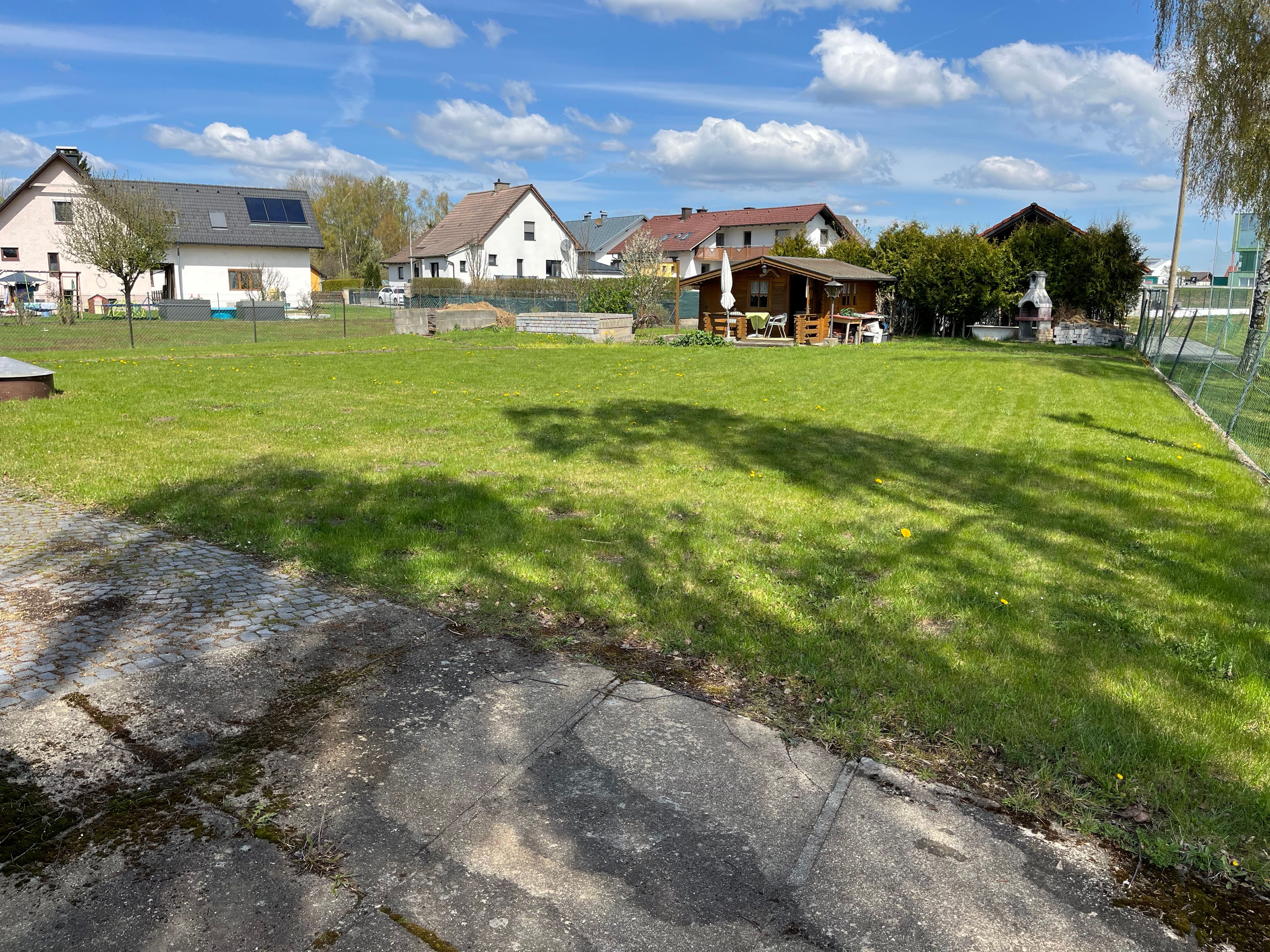 Ein Garten mit Rasenfläche, einem Gartenhaus und einem Grillplatz im Hintergrund. Mehrere Wohnhäuser sind im Hintergrund zu sehen.