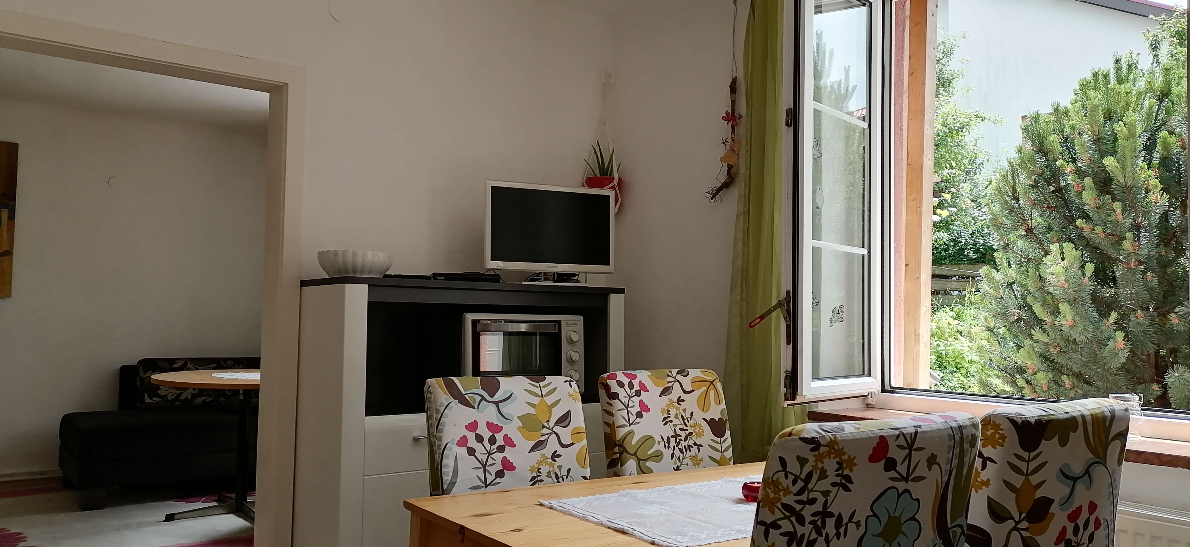 Essbereich mit Tisch, bunten Stühlen, Fernseher und offenem Fenster mit Blick ins Grüne.