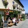 Eine Gruppe von Menschen sitzt vor einem Gasthof an Holztischen auf einer gepflasterten Straße.