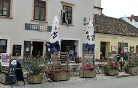 Außenansicht eines Cafés mit Tischen, Stühlen und Sonnenschirmen vor dem Gebäude.