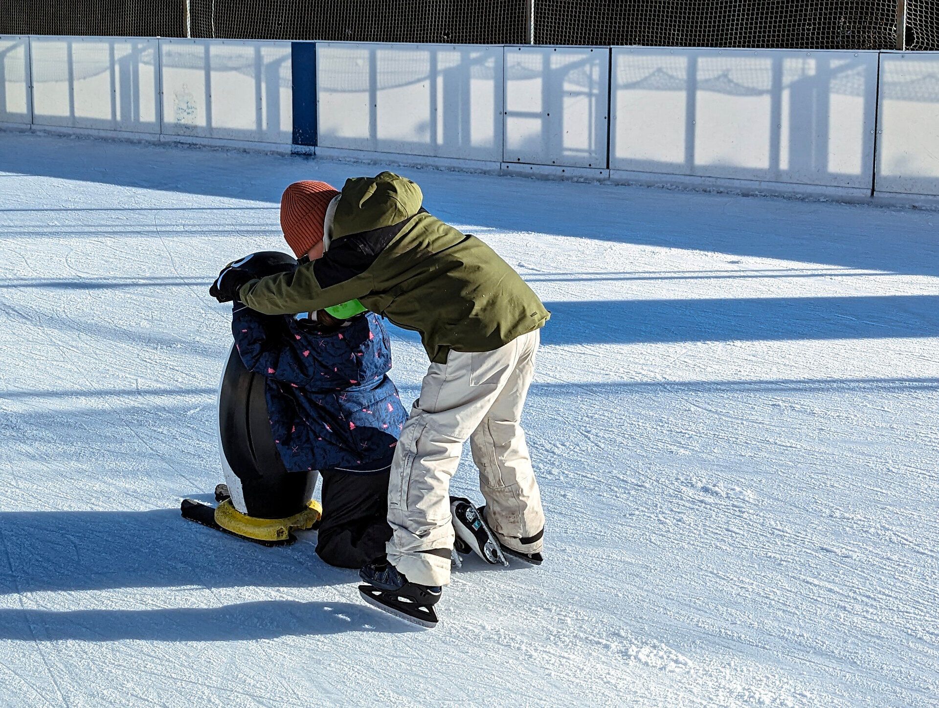 Auf der glitzernden Eisfläche gleiten Kinder voller Freude und Begeisterung. Mit einem stabilisierenden Pinguin als Unterstützung lernen sie spielerisch das Eislaufen und genießen die frische Winterluft. Lachen und fröhliche Rufe erfüllen die Atmosphäre, während die Sonne sanft auf das Eis scheint.