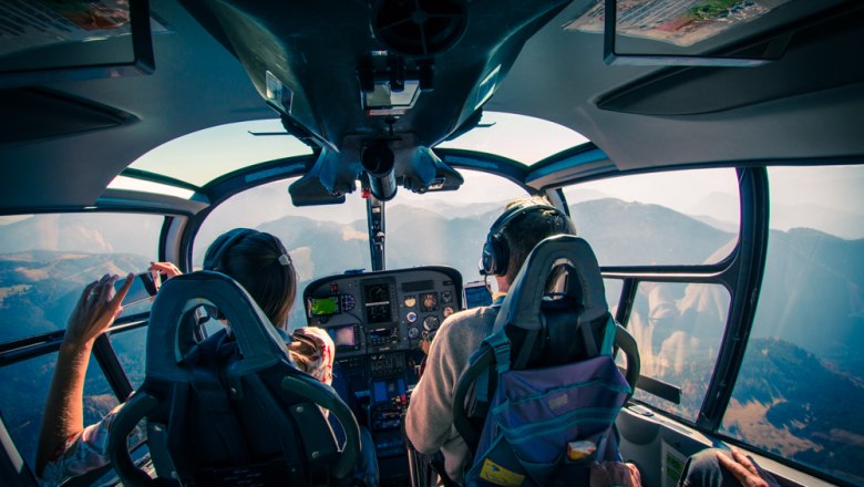 Blick aus dem Cockpit eines Helikopters mit zwei Personen, die über eine Berglandschaft fliegen.