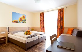Ein helles G&auml;stezimmer mit zwei Betten, einem Tisch, einem Sofa und einem Bild von Sonnenblumen an der Wand.