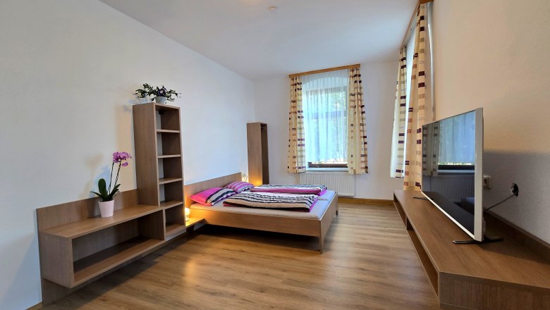 Modernes Schlafzimmer mit Holzboden, Doppelbett, Fernseher und Regalen. Große Fenster mit gestreiften Vorhängen.