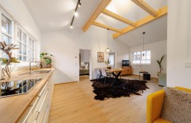 Modernes Wohnzimmer mit Holzbalken, Esstisch, gelbem Sessel und offener Küche.