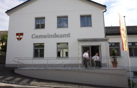 Ein modernes Gemeindeamt mit weißer Fassade, zwei Personen stehen auf einer Rampe, daneben weht eine Flagge.
