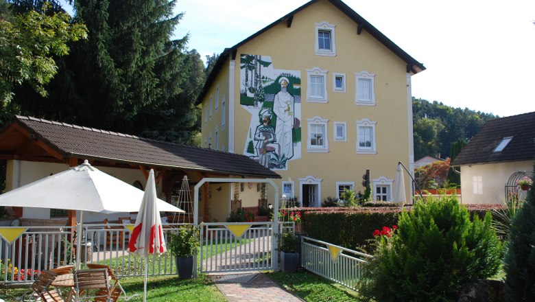 Gelbes Gebäude mit Wandgemälde, Garten mit Sonnenschirm und Sitzbereich.