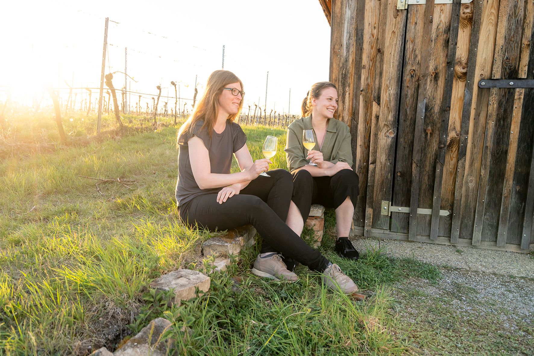 Zwei Frauen sitzen mit Weingläsern vor einer Holzhütte im Weinberg.
