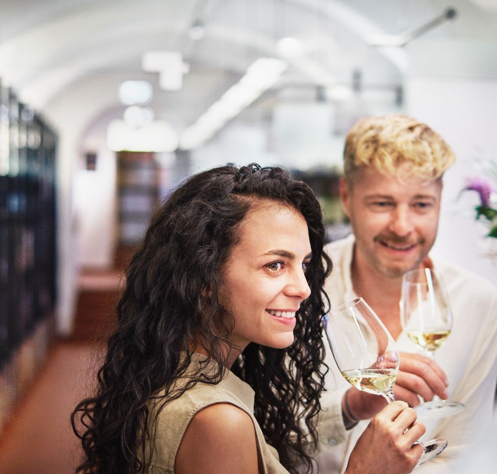 Zwei Personen stehen in einer Vinothek und werden von einer Person beraten. Regale mit Weinflaschen sind im Hintergrund zu sehen.