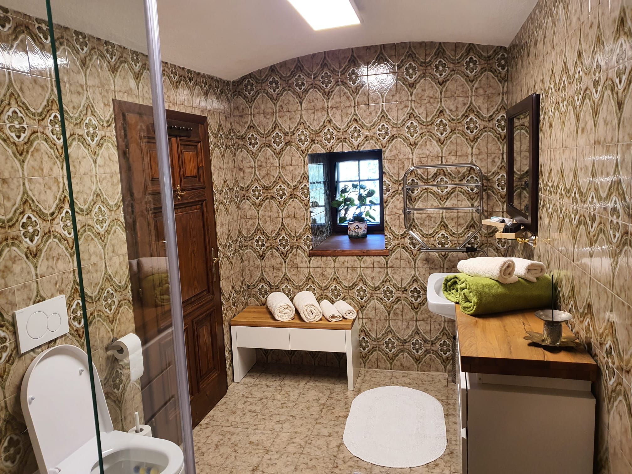 Ein Badezimmer mit gemusterten Fliesen, einem Fenster, einer Toilette, einem Waschbecken und Handtüchern auf einer Holzablage.