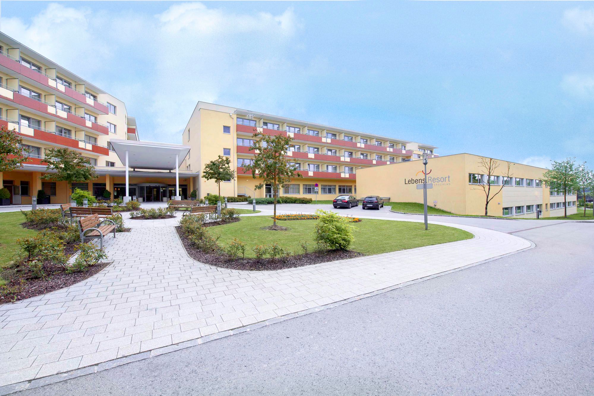 Außenansicht eines modernen Gebäudekomplexes mit gepflegtem Garten und Parkplätzen.