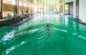 Person schwimmt in einem Hallenbad mit grünen Wasser und Liegestühlen am Rand.