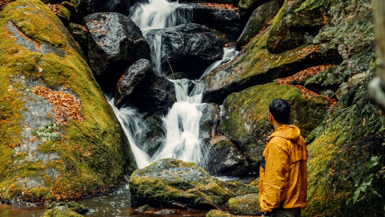 Ein Mann in gelber Jacke steht vor einem kleinen Wasserfall im Wald.