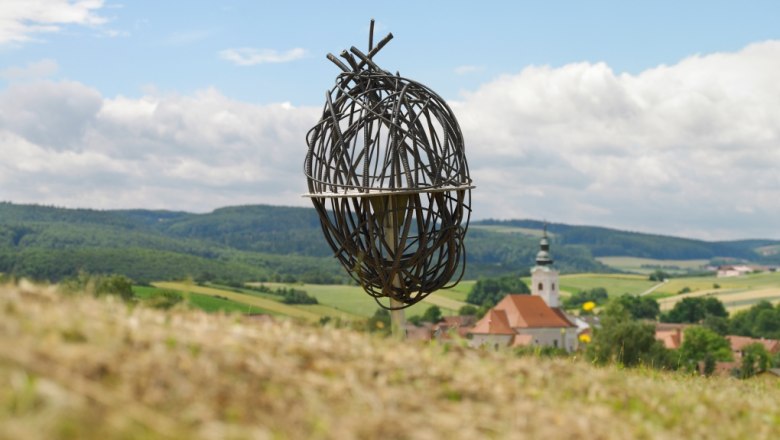 Kunstinstallation aus Metall in einer ländlichen Landschaft mit Kirche im Hintergrund.