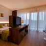 Modernes Hotelzimmer mit Doppelbett, Fernseher, Schreibtisch und großen Fenstern mit Vorhängen.