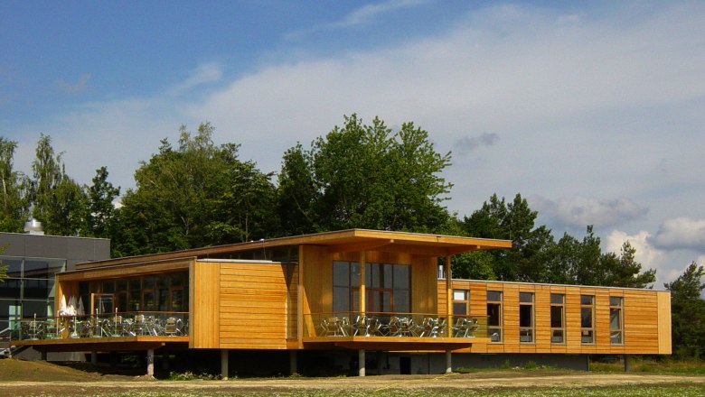 Modernes Gebäude aus Holz mit großen Fenstern und Terrasse, umgeben von Bäumen.