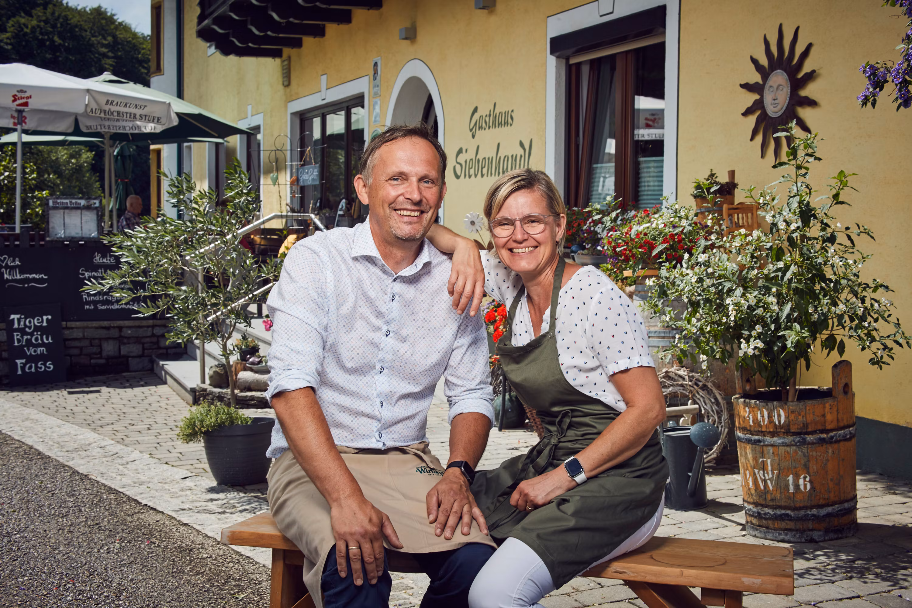 Lächelndes Paar vor Gasthaus Siebenhandl, umgeben von Pflanzen und Dekoration.