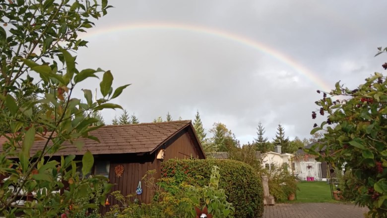Ein Regenbogen spannt sich über einem Garten mit einem Holzhäuschen und Pflanzen.