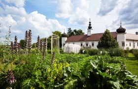 Ein Klostergebäude mit Zwiebeltürmen hinter einem üppigen Garten unter blauem Himmel.